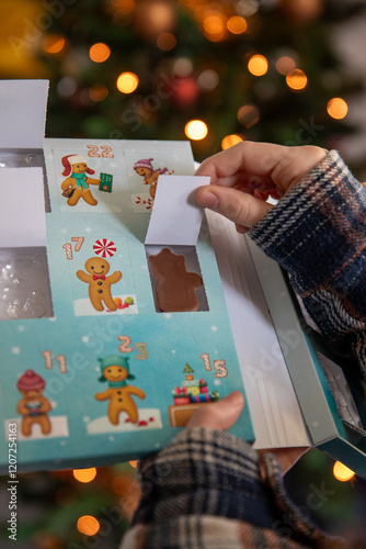 Gros plan d'un enfant ouvrant la fenêtre d'un calendrier de l'Avent avec un chocolat, sapin de noël avec guirlande lumineuse en fond.