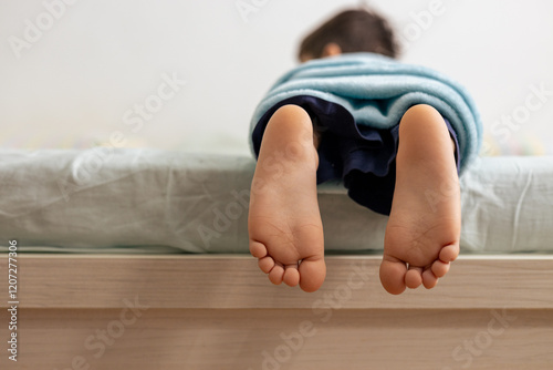 Child covered with light blue blanket lying on bed with feet dangling off edge