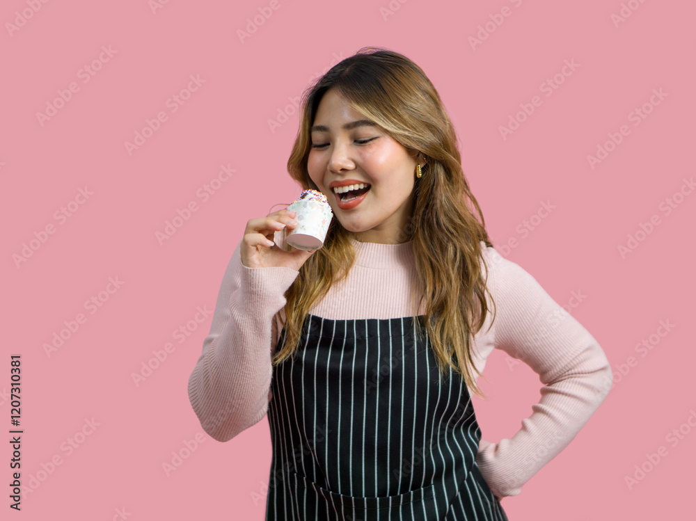 Young asian woman with an apron in celebratory or festive mood holding cupcake, decorated with white frosting and chocolate chip. Portrait on pink background with studio light, Isolated. Young asian woman with an apron in celebratory or festive mood holding cupcake, decorated with white frosting and chocolate chip. Portrait on pink background with studio light, Isolated.