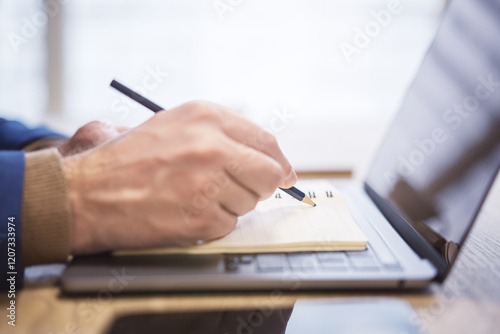 In close view, a man's hand writing down notes in a notepad on a laptop, against a hazy backdrop photo