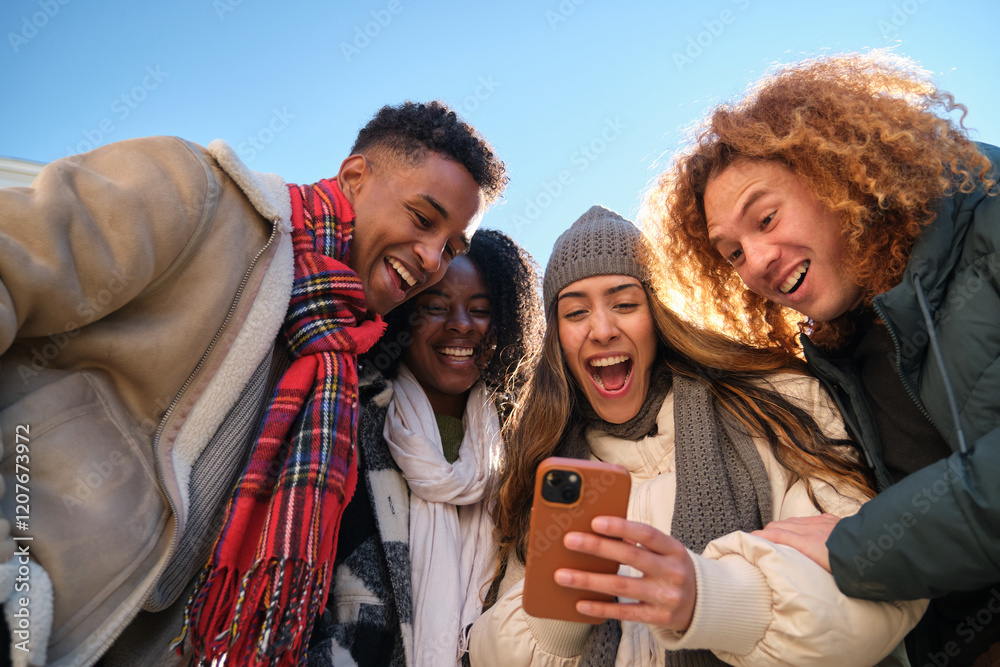 Four young cheerful multi ethnic people having fun together outdoors in winter, looking at smartphone screen