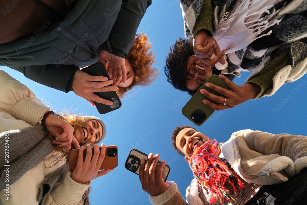 Four cheerful young people using their smartphones, forming a circle, low angle view against a blue sky