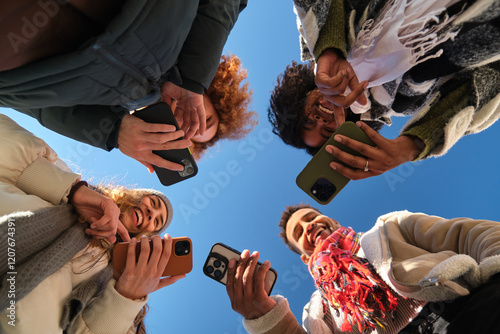 Obraz na płótnie Four cheerful young people using their smartphones, forming a circle, low angle view against a blue sky w salonie