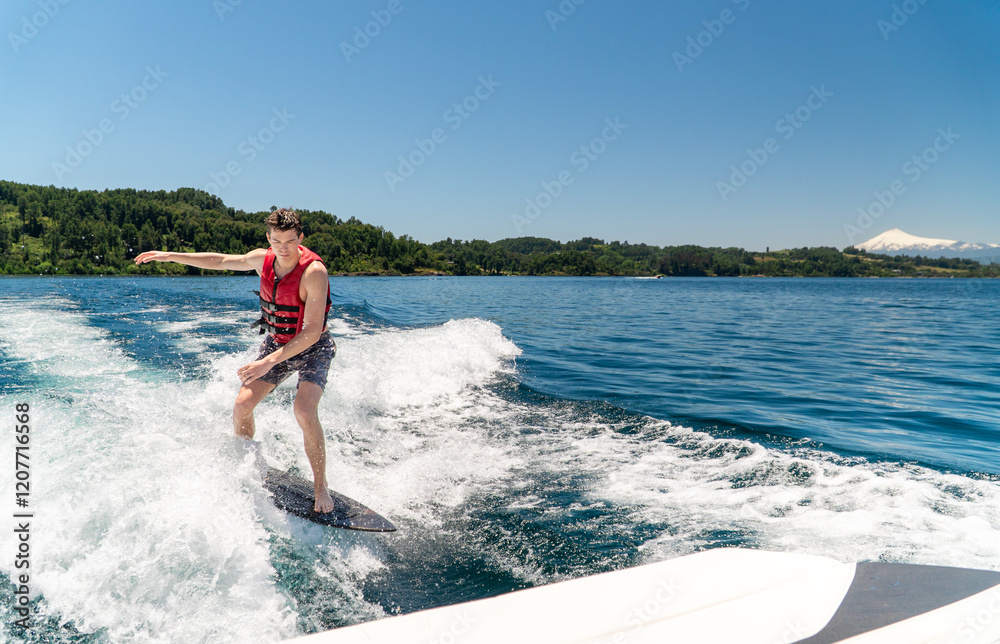 Young male wearing a red life jacket wake surfing wave behind boat at a lake with a big volcano on the back.