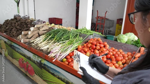 An employee of a fruit and vegetable supermarket is taking inventory of some fruits such as tomatoes, carrots and onions with a spreadsheet and a pen is writing down the quantities.