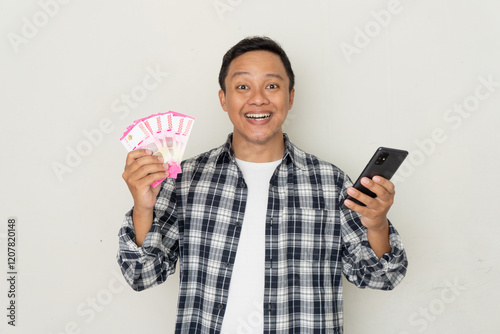 Young Asian man excited and happy while holding mobile phone and bunch of money in Rupiah currency. photo