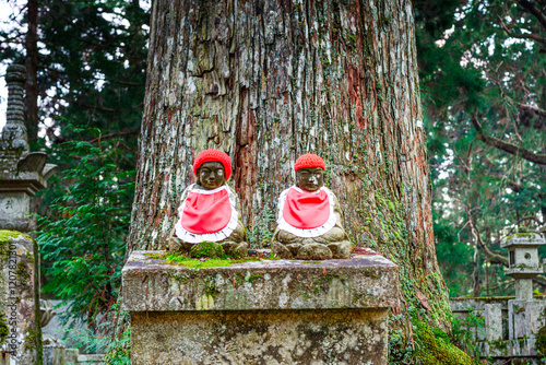 Two red hat Jizo statues under old tree trunk. Buddhist cemetery of Oku-no-in, Koyasan (Koya-san), Kansai, Honshu, Japan photo