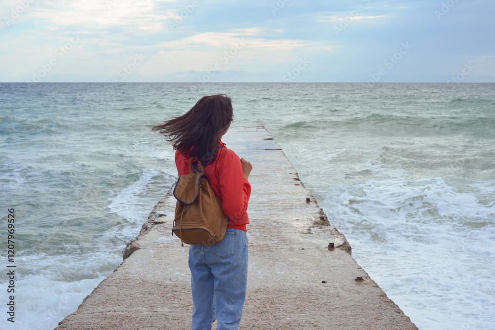 Rear view of a young woman tourist with a backpack standing on the pier and waves hitting the pier in a storm. City of Sudak, Crimea.
