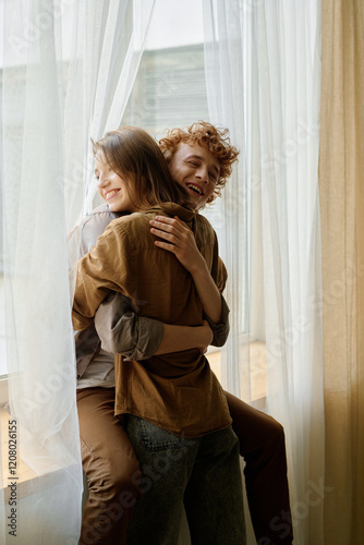 Vertical shot of red haired man sitting on windowsill smiling and hugging his beloved beautiful wife, they illumed by sunray photo