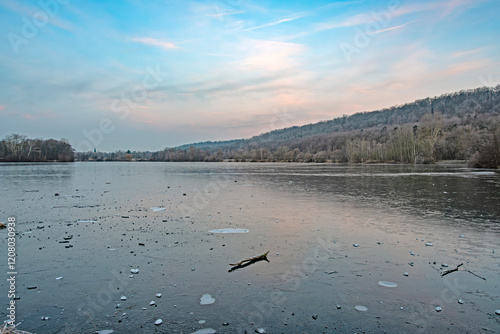 Gefrorener See bis fast zum Horizont, am linken Bildrand ein mit Bäumen bewachsener Hügel der sich sanft am See in die Höhe erstreckt. Über der Szene liegt ein winterlicher hell blau-weißer Himmel photo