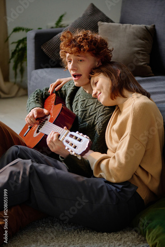 Attractive woman hugging her boyfriend who playing guitar and signing, they sitting on fluffy carpet and leaning on sofa