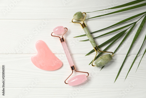 Face rollers, gua sha tool and leaf on white wooden background, flat lay photo