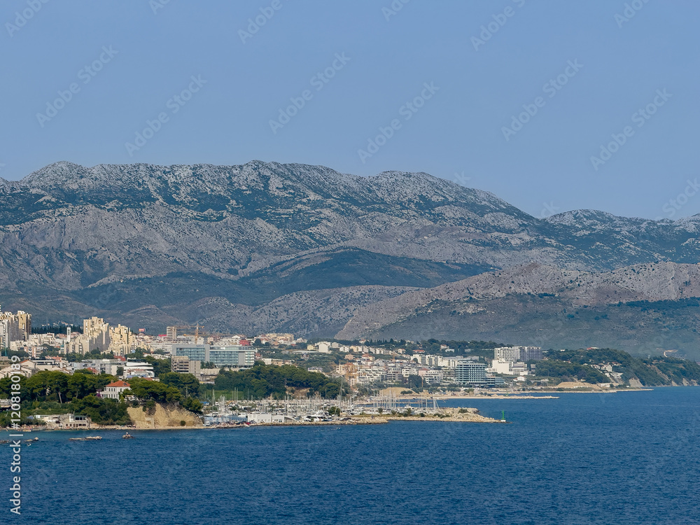 Obraz premium Split, Croatia - July 1, 2024: Zenta Marina yacht harbor in center of large landscape showing tall mountain range on horizon and an urban jungle around it. Tall apartment buildings and tourism-related