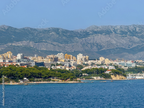 Split, Croatia - July 1, 2024: Coastline west of Marina Zenta Yacht port. Green trees above yellow rocks, beaches all the way with tall buildings cityscape in back in front of gray-green large mountai photo