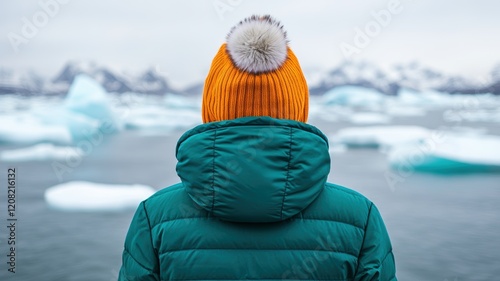 A person wearing a colorful knitted hat stands by icy waters, gazing at the serene landscape filled with floating icebergs.