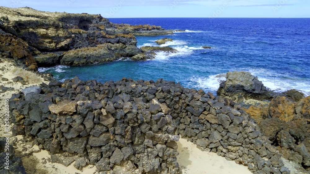 Volcanic rocks are forming a wall in Charco del Palo, Lanzarote, Canary Islands, Spain, near a beautiful blue sea with waves crashing on the rocky shore Volcanic rocks are forming a wall in Charco del Palo, Lanzarote, Canary Islands, Spain, near a beautiful blue sea with waves crashing on the rocky shore