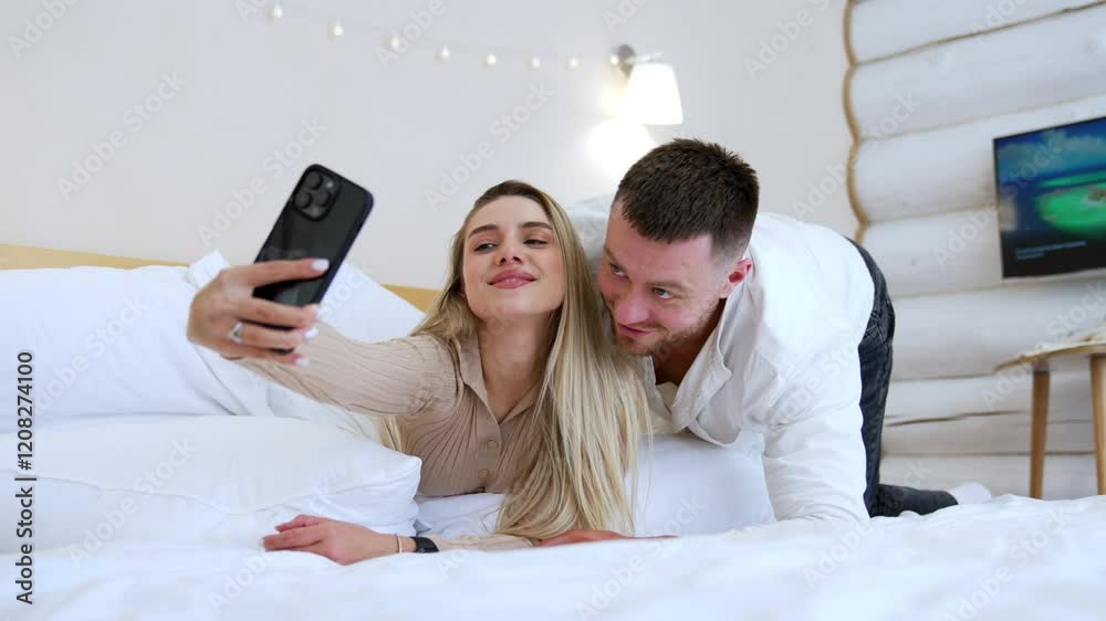Beautiful blonde woman lies on the bed taking selfie. Bearded man lies beside looking at telephone camera. Beautiful blonde woman lies on the bed taking selfie. Bearded man lies beside looking at telephone camera.