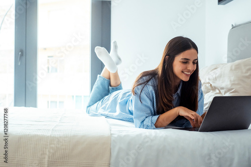 One laxy and cute young woman chatting online at home laying on bed with a laptop. Morning light in background. Concept of indoor leisure activity with computer or smart working photo