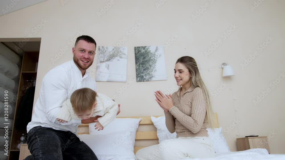 Bearded Caucasian man holds and tosses his little baby son. Happy lovely child is laughing adorably. Mother sitting beside claps hands. Low angle view. Bearded Caucasian man holds and tosses his little baby son. Happy lovely child is laughing adorably. Mother sitting beside claps hands. Low angle view.
