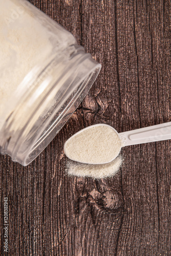Plastic jar with powder and measuring spoon. Protein or collagen on wooden background. Scattered powder. Vertical