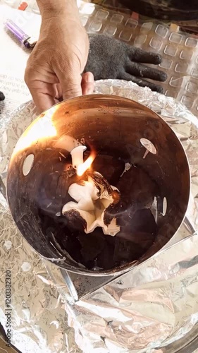 A person uses a candle to set fire to a piece of paper in a charcoal container of a smoking barrel.