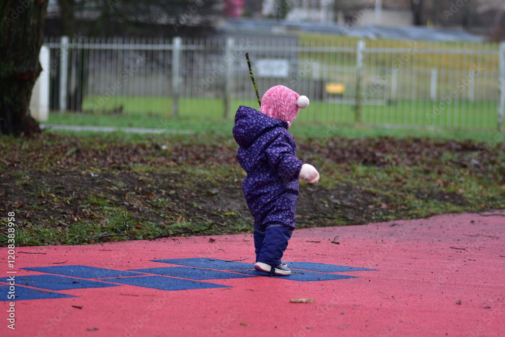 Obraz premium Baby girl standing backwards and holding tree branch in children playground in cold weather autumn winter