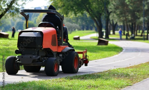 Big professional riding lawn mower with an operator on it driving in city park in sunny summer day.
