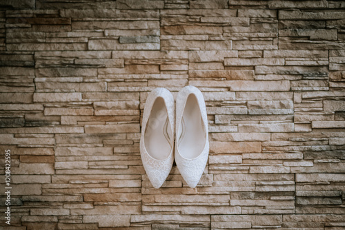 A pair of white lace bridal shoes neatly arranged on a textured stone wall backdrop, creating a stylish and elegant composition. photo