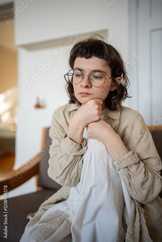 Thoughtful bored lonely teen girl in glasses feeling apathy sitting on couch at home. Teenager suffer from depression, mental disorder, anhedonia, stress. Burnout syndrome after stress life period. photo