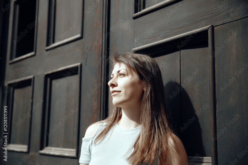Fototapeta premium Thoughtful woman standing by a rustic wooden door under natural sunlight