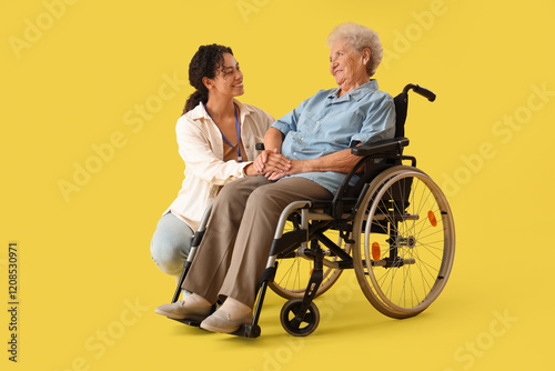 Young African-American female medical worker with elderly woman on wheelchair against yellow background