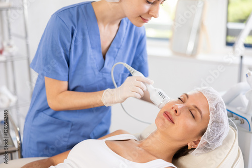 Young adult female patient receiving professional hardware facial procedure at beauty clinic using cooling attachment to alleviate inflammation, rejuvenate of skin and enhance complexion