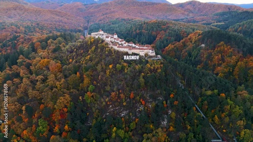 Dynamic aerial shot circling the Râșnov Fortress, highlighting its sturdy walls, rustic charm, and breathtaking backdrop of the Carpathian Mountains.
