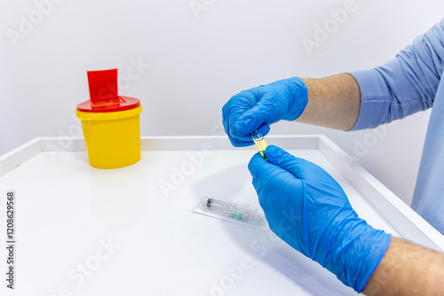 On a white background, hands in blue latex gloves remove and break an ampoule, drawing medication into a syringe. Hospital or emergency room scene. Photo of needle, syringe and medication ampoule.