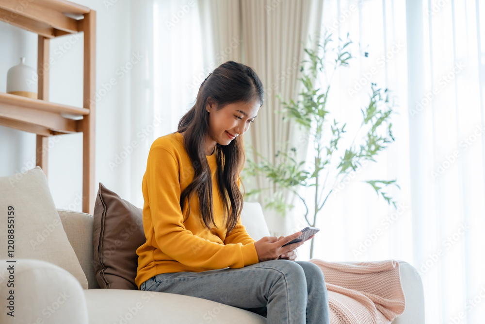 Smiling pretty young Asian woman sitting on cozy couch, using funny mobile apps in living room. Woman at home, Doing online shopping, Messaging friends, Posting on social Media Smiling pretty young Asian woman sitting on cozy couch, using funny mobile apps in living room. Woman at home, Doing online shopping, Messaging friends, Posting on social Media