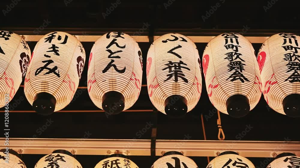 A row of lanterns with Japanese writing on them on street stand shop. The lanterns are lit up and hanging from the ceiling