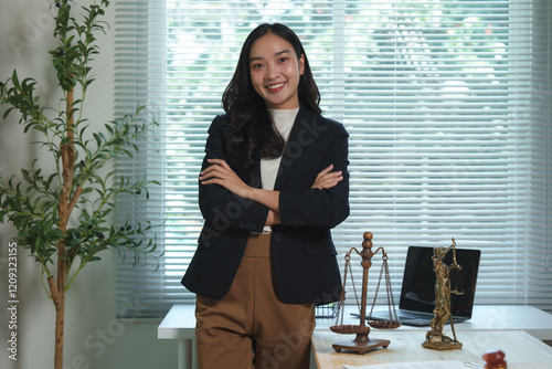 Young female lawyer standing confidently in her office with arms crossed, surrounded by legal symbols like the themis statue and scales of justice, embodying integrity and fairness photo