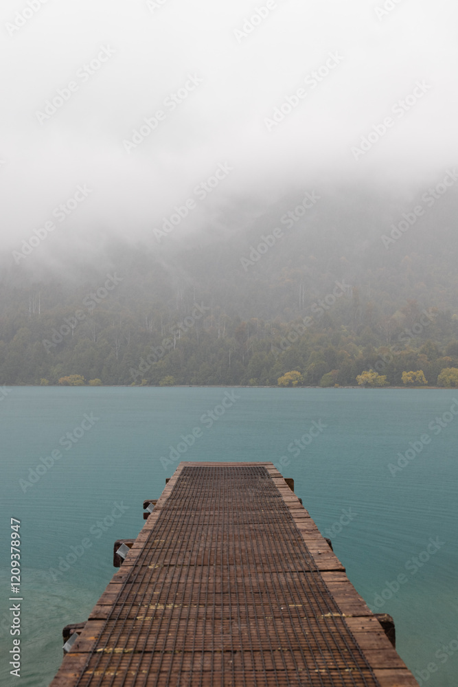 Obraz premium Small jetty towards a lake with fog-covered mountain range and turquoise water (South Island, New Zealand)