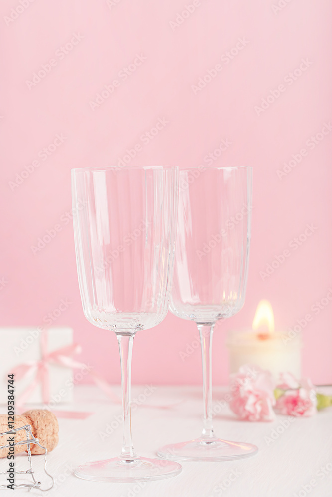 Two empty transparent wine glasses served on white wooden table with glowing candle light, champagne bottle top, flowers and gift box against pink background with copy space prepared for celebration Two empty transparent wine glasses served on white wooden table with glowing candle light, champagne bottle top, flowers and gift box against pink background with copy space prepared for celebration