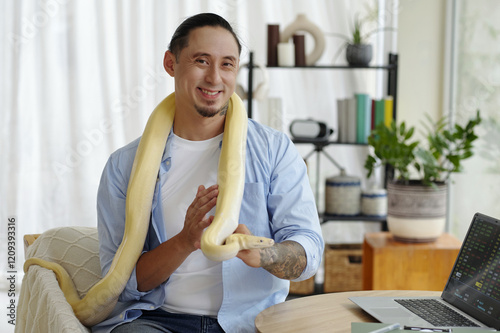 Portrait of man smiling while sitting indoors with yellow snake draped on shoulders, displaying ease and comfort with pet snake, near modern home office setup