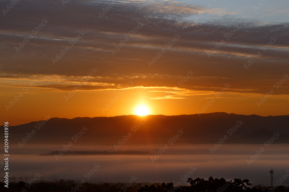 Obraz premium Sunrise over Misty Mountains, A breathtaking sunrise paints the sky in vibrant hues of orange and gold as the sun rises majestically behind a range of misty mountains.