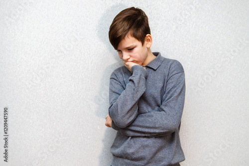 A ten year old boy in a gray sweater stands by the wall. Sadly put his head on his hand and looks down photo