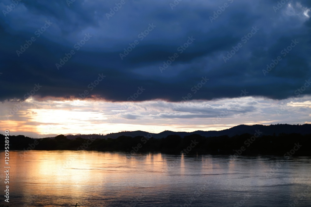 Dramatic sunset over a wide river with dark storm clouds gathering above silhouetted hills..Concept: Moody landscape, storm approaching, dramatic sunset, nature's power. Dramatic sunset over a wide river with dark storm clouds gathering above silhouetted hills..Concept: Moody landscape, storm approaching, dramatic sunset, nature's power.