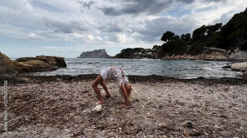 Slow motion sequence of a young girl performing a back walkover on the beach. On the third try, she manages to get back to her original position.