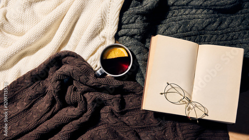 Aerial view of open book and glasses resting on knitted blankets with hot drink in metal mug with orange slice, representing winter relaxation and creative inspiration. Concept of style, aesthetic. photo