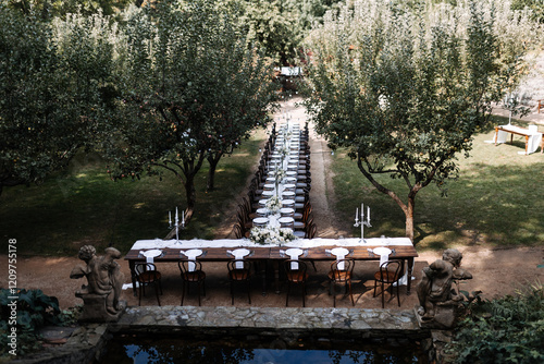 Long banquet table set for an outdoor event in a garden surrounded by apple trees. Elegant dining setup with white tableware, floral arrangements, and candelabras