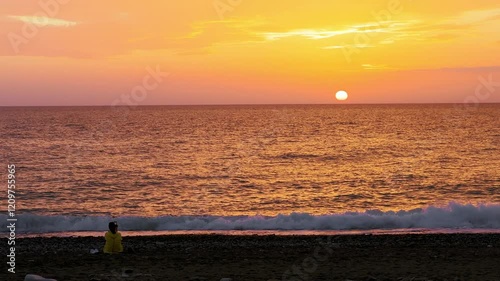 Woman sits on seashore near waves washing shore, films sunset on mobile phone camera. Tourist girl takes photographs or videos of seascape with evening sky, dressed in yellow t-shirt