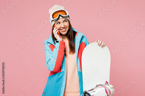 Snowboarder young woman wear warm blue padded jacket ski goggles mask spend extreme weekend winter season in mountains hold snowboard talk speak on mobile cell phone isolated on plain pink background. photo