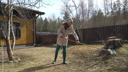 A woman in outdoor clothing raking leaves and dead grass clearing a garden in early spring. The scene with natural spring sunlight, seasonal yard work and connection with nature.