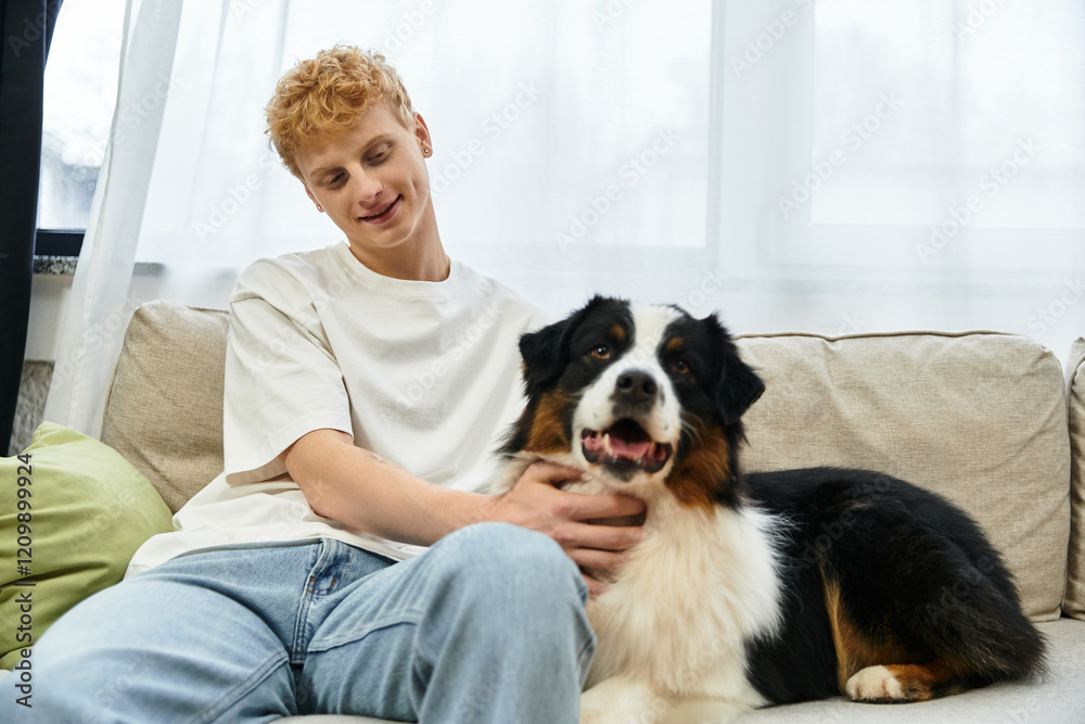 A young redhead man relaxes on a couch, lovingly petting his Australian Shepherd dog at home. A young redhead man relaxes on a couch, lovingly petting his Australian Shepherd dog at home.